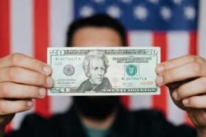 Close-up of a man holding a 20-dollar bill with an American flag blurred in the background, symbolizing finance and patriotism.